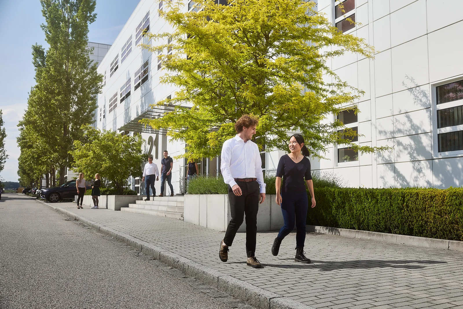 Zwei Personen gehen auf einem Weg und unterhalten sich. Im Hintergrund ist ein DRÄXLMAIER Gebäude und Bäume.