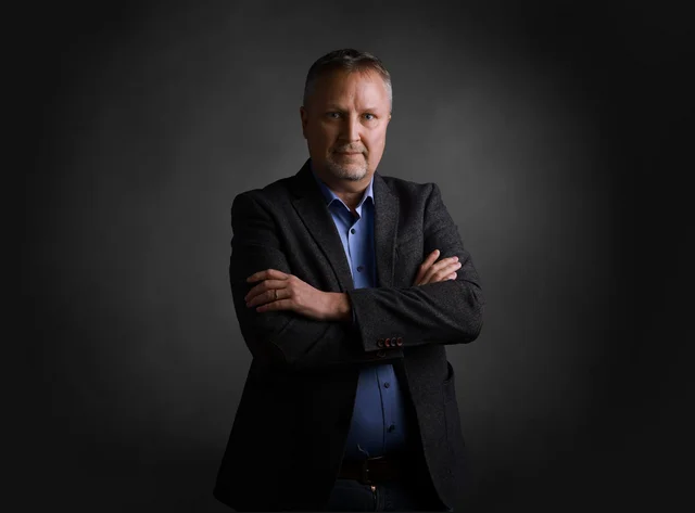Wolfgang Ein Mann steht lächelnd mit verschränkten Armen vor schwarzem Hintergrund.
