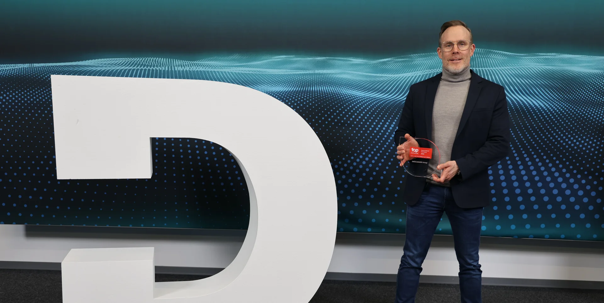 Ein großes weißes DRÄXLMAIER Logo. Rechts daneben steht ein Mann mit dem Top Employer Award.