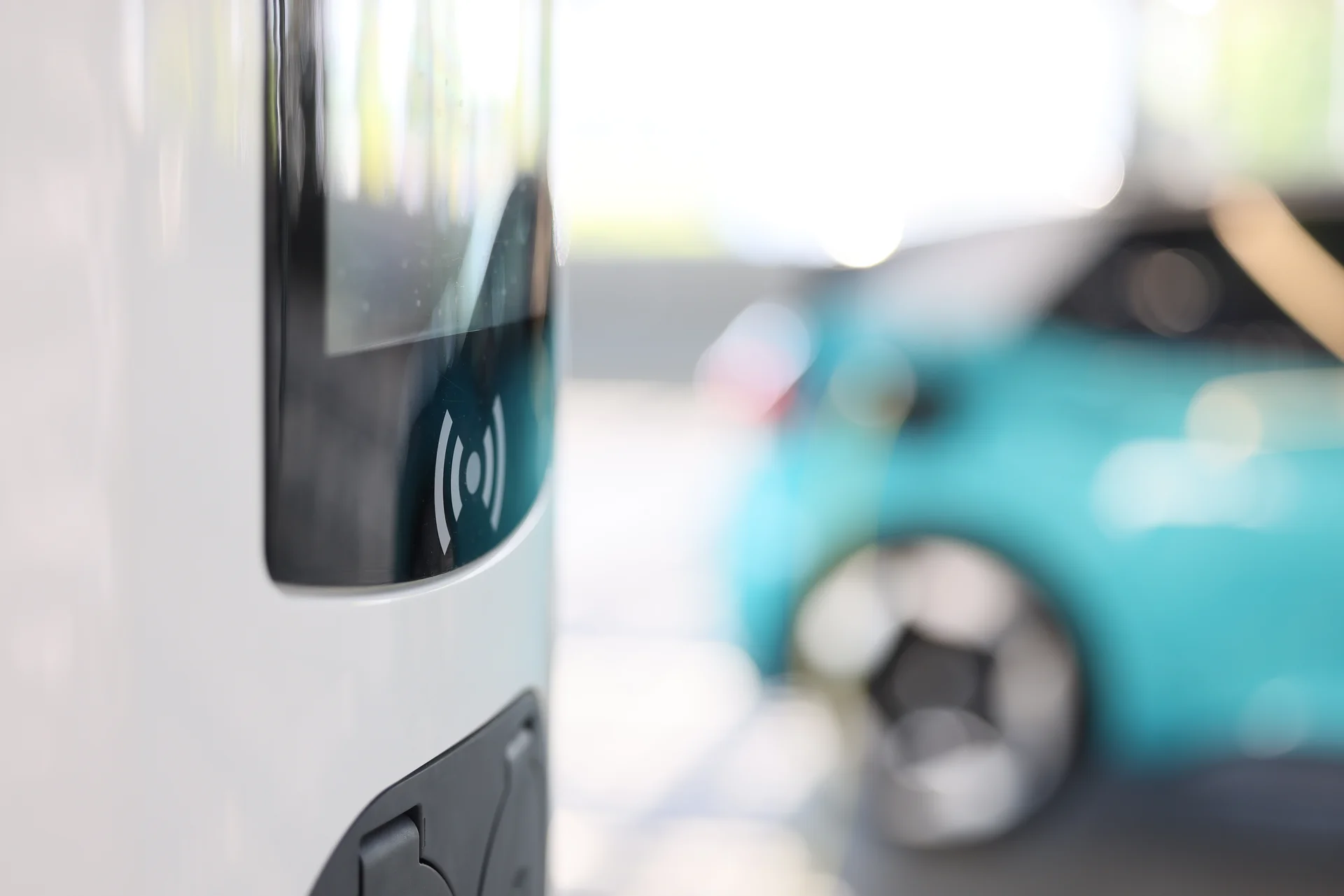 Im Vordergrund ist eine E-Ladesäule. Im Hintergrund sieht man ein türkises Auto.