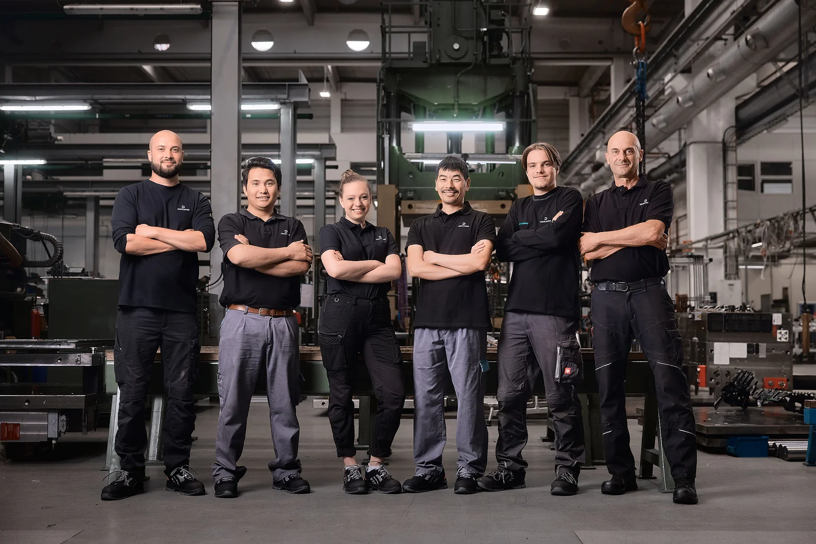 Six DRÄXLMAIER production employees stand in a row in a hall. They smile into the camera.