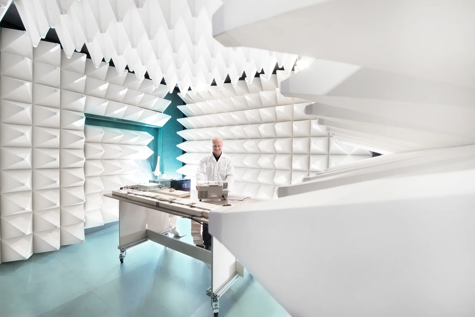 A person stands in an anechoic chamber, surrounded by white foam wedges, and works at a table with electronic devices.