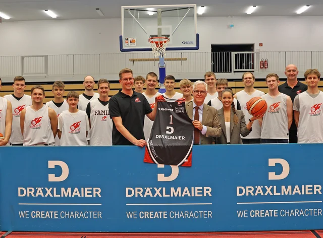 Die Baskets Vilsbiburg Die Baskets Vilsbiburg und der Head of Marketing der Firma DRÄXLMAIER stehen lächlend in der Basketballhalle.