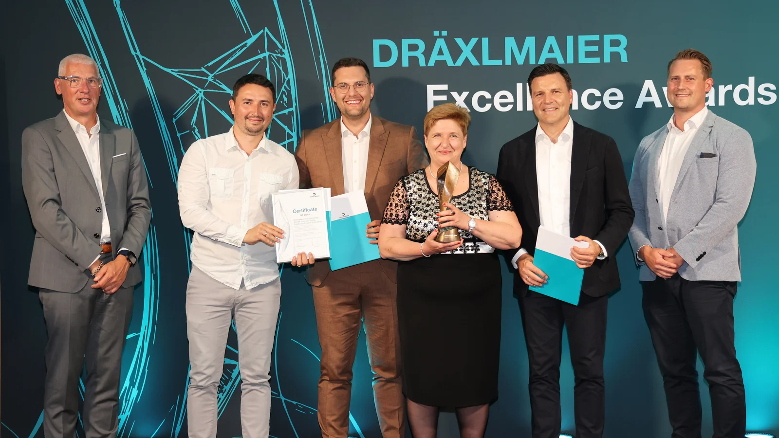 6 Leute stehen vor einer Leinwand mit der Aufschrift "DRÄXLMAIER Excellence Awards". Sie halten Urkunden und eine Trophäe.