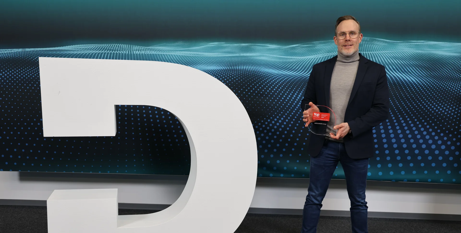 A large white DRÄXLMAIER logo. To the right is a man with the Top Employer Award.