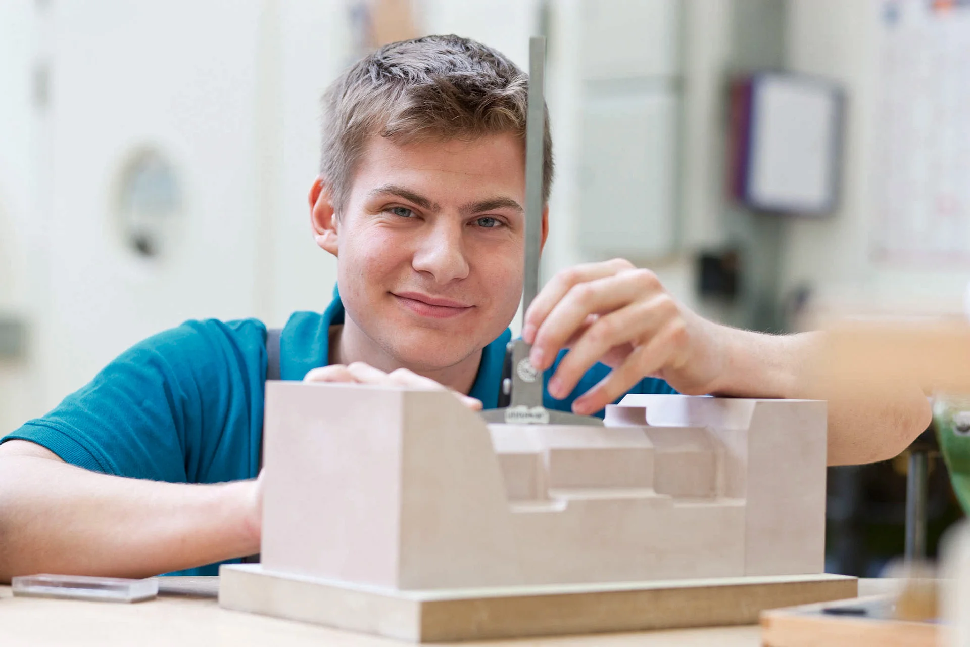 Ein Auszubildender bei DRÄXLMAIER in Vilsbiburg lächelt in die Kamera, und hält ein Werkstück in der Hand.