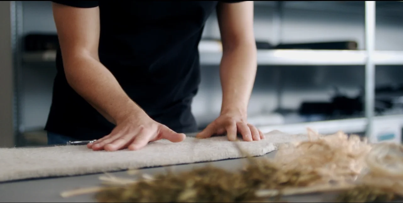 A person works a natural fibre mat by hand on a workbench.
