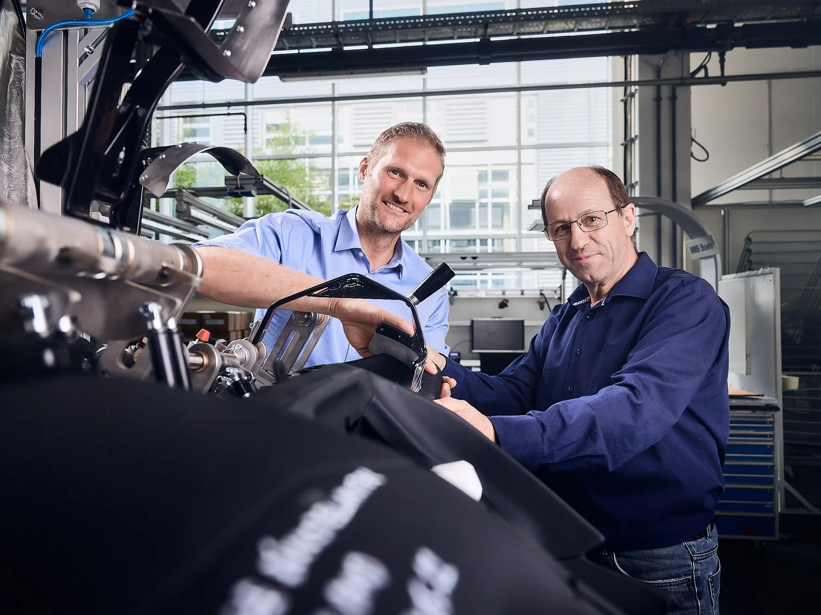 Two men in shirts smile at the camera and hold a component together.